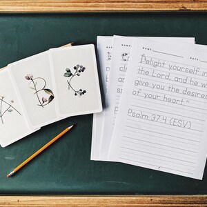 May include: A wooden-framed chalkboard displays alphabet cards with floral illustrations and a pencil. The cards spell out "ABC" with botanical designs. Beside them are lined papers with the text "Delight yourself in the Lord, and he will give you the desires of your heart." Psalm 37:4 (ESV).