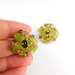 May include: A pair of handmade stud earrings. Each earring features a central dark green gemstone surrounded by light green and gold-colored beads. The earrings have a floral design and are set against a white background.