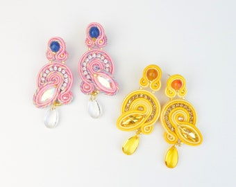 May include: Two pairs of handmade earrings. One pair is pink with blue and clear accents, the other is yellow with orange and clear accents. Both pairs feature intricate beadwork and dangling crystal-like teardrops.