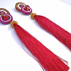 May include: A pair of long, red tassel earrings with a pink and white beaded design and a red rhinestone in the center.
