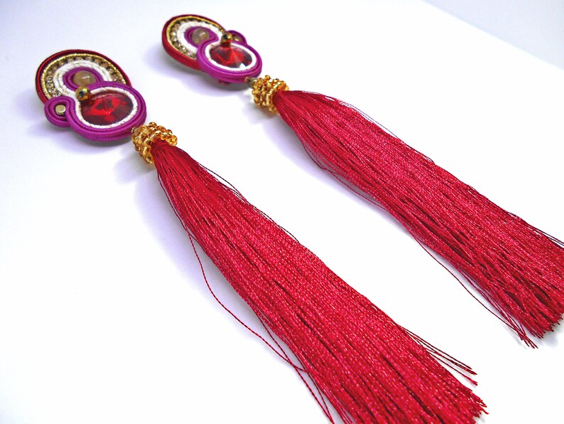 May include: A pair of long, red tassel earrings with a pink and white beaded design and a red rhinestone in the center.