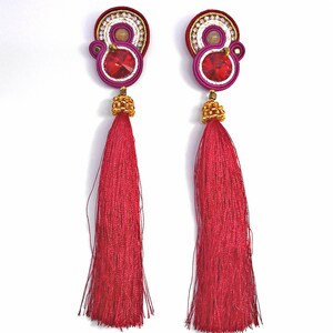 May include: A pair of long, red tassel earrings with a pink and white beaded design and a red crystal in the center. The earrings are handmade and feature a gold-tone finish.