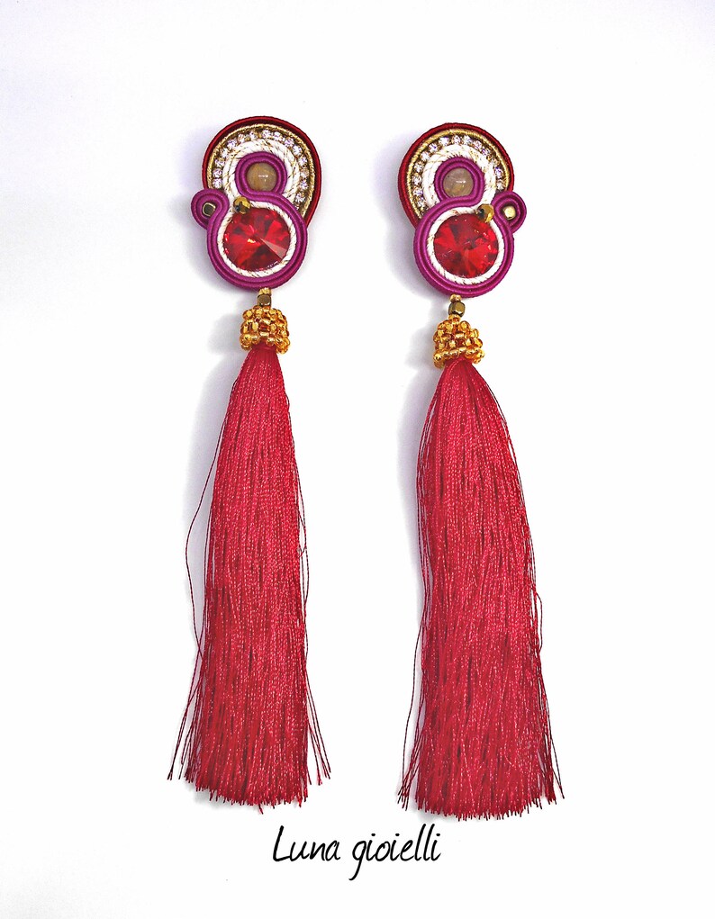 May include: A pair of long, red tassel earrings with a pink and white beaded design and a red crystal in the center. The earrings are handmade and feature a gold-tone finish.
