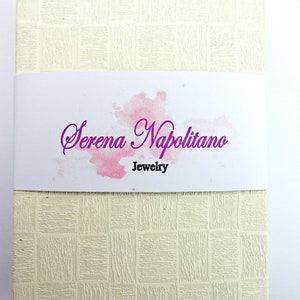 May include: A white box with a textured surface and a pink watercolor design. The text "Serena Napolitano Jewelry" is printed in purple on the box.