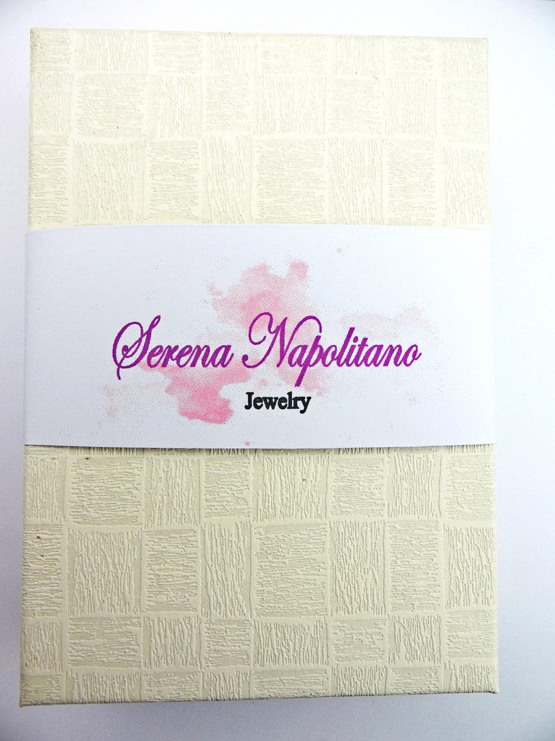 May include: A white box with a textured surface and a pink watercolor design. The text "Serena Napolitano Jewelry" is printed in purple on the box.