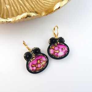 Peut inclure: Une paire de boucles d'oreilles rose et or avec des accents noirs. Les boucles d'oreilles sont faites d'un tissu tissé avec un motif rose et or. Les boucles d'oreilles sont suspendues à des crochets dorés.