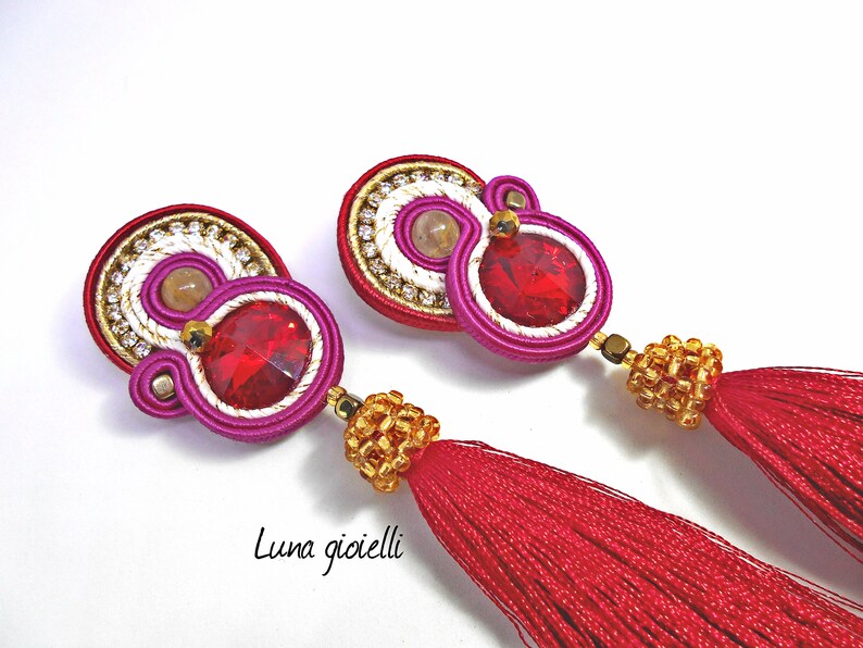 May include: A pair of red tassel earrings with a pink, white, and gold beaded design. The earrings feature a red crystal and a small, round, beige stone.