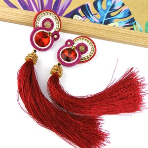 May include: A pair of red tassel earrings with gold and white accents. The earrings feature a red, white, and gold circular design with a red crystal in the center.
