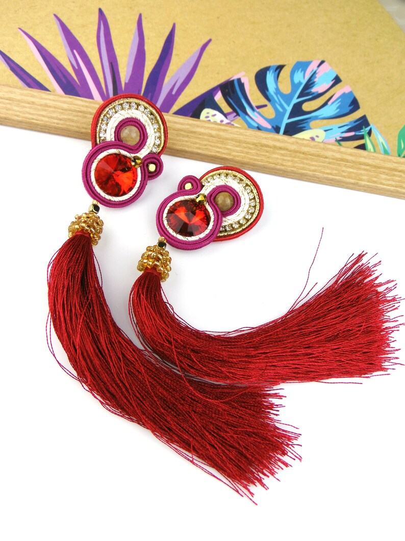 May include: A pair of red tassel earrings with gold and white accents. The earrings feature a red, white, and gold circular design with a red crystal in the center.