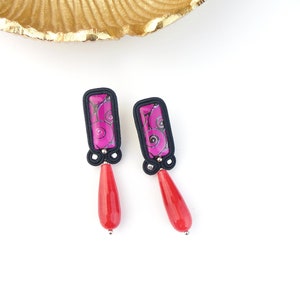 May include: A pair of dangle earrings with black and pink patterned rectangular charms and red teardrop-shaped beads.
