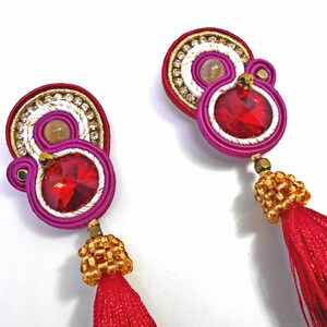 May include: A pair of earrings with red and pink tassels. The earrings feature a gold and white beaded design with a red rhinestone in the center.