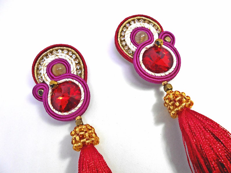 May include: A pair of earrings with red and pink tassels. The earrings feature a gold and white beaded design with a red rhinestone in the center.
