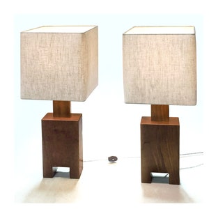 May include: Two matching wooden table lamps. Each lamp has a square, beige fabric shade atop a rectangular wooden base. The base is a dark brown color, with a lighter brown wooden support connecting the base to the shade. The lamps are on a white surface.