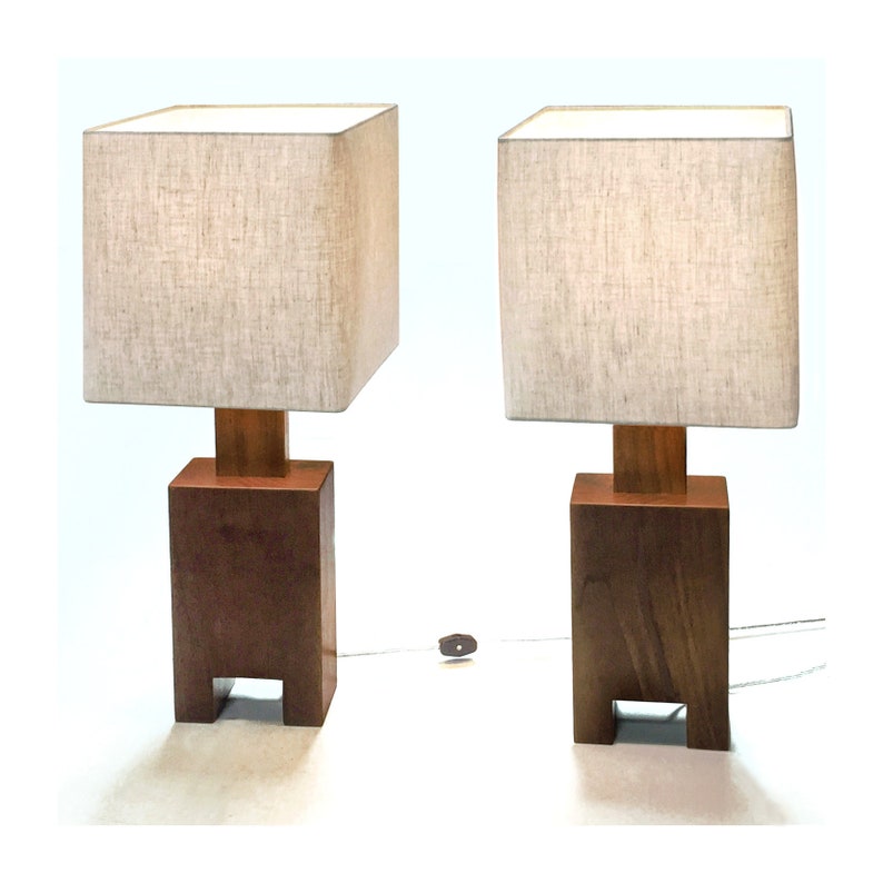 May include: Two matching wooden table lamps. Each lamp has a square, beige fabric shade atop a rectangular wooden base. The base is a dark brown color, with a lighter brown wooden support connecting the base to the shade. The lamps are on a white surface.
