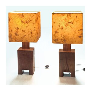 May include: Two matching wooden table lamps. Each lamp has a square, textured, amber-colored shade atop a wooden base. The base is a dark brown, rectangular block with a smaller square wooden support. The lamps are connected to a power source.
