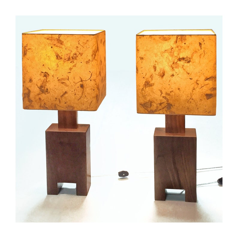 May include: Two matching wooden table lamps. Each lamp has a square, textured, amber-colored shade atop a wooden base. The base is a dark brown, rectangular block with a smaller square wooden support. The lamps are connected to a power source.
