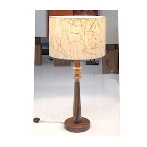 May include: A wooden table lamp with a cylindrical lampshade. The lampshade is beige with a textured pattern. The lamp base is dark brown, with a round base and a conical stem. The lamp has a decorative section with three stacked rings. The lamp is plugged into a wall outlet.