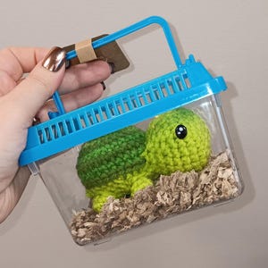 May include: A crocheted green turtle with black eyes inside a clear plastic carrier with a blue handle. The carrier contains wood shavings. The turtle is a handmade item.