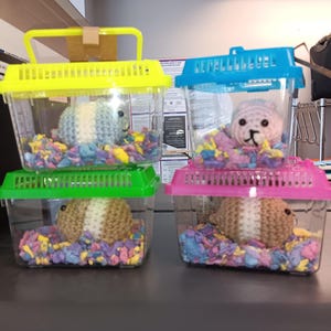 May include: Four clear plastic pet carriers with colorful lids in yellow, green, blue, and pink. Each carrier contains a crocheted animal and colorful paper bedding. The animals are in shades of blue, pink, and brown.