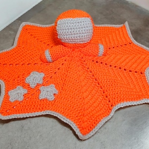 May include: A crocheted orange star-shaped baby blanket with a gray crocheted astronaut toy in the center. The blanket has three gray crocheted stars on it.