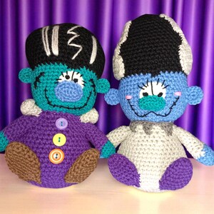 May include: Two handmade crochet dolls, resembling Frankenstein's monster and his bride, sit side-by-side. The dolls have blue faces, black hair, and are wearing purple and white outfits. The dolls are on a white surface with a purple background.