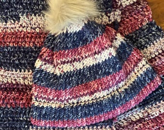 May include: A striped knit hat with a fluffy pom pom and a matching striped scarf. The hat and scarf are in shades of pink, purple, and blue.