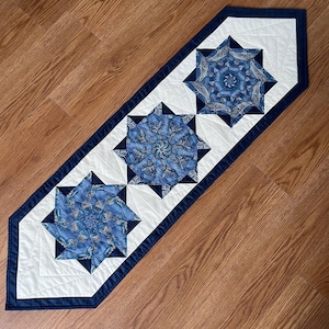 May include: A blue and white table runner with a geometric pattern. The runner has a navy blue border and three large, blue star-shaped designs.