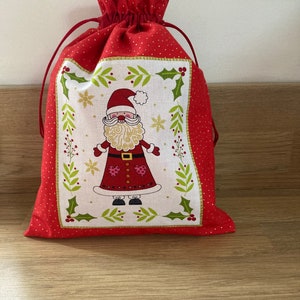 Peut inclure: Sac cadeau de Noël rouge avec fermeture à cordon. Le sac présente un panneau central avec une illustration du Père Noël entourée de houx et de flocons de neige. Le tissu rouge du sac a un motif à pois dorés.