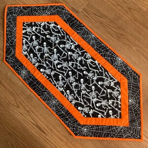 May include: Halloween table runner with a black background and white spiderwebs on the edges. The centre has a black fabric with white skeleton figures. The runner is bordered with orange fabric.
