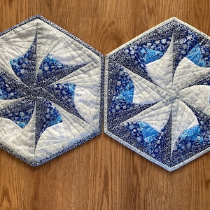 May include: Two hexagonal quilted placemats featuring a snowflake design. The placemats are made from white fabric with blue and silver accents. The snowflake design is created using a series of overlapping fabric pieces.