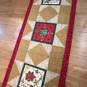 May include: A red and gold Christmas table runner with a white star pattern and three green and red floral squares. The runner has a red border with white polka dots.