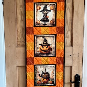 May include: A Halloween wall hanging with three illustrated panels. The top panel depicts a witch, the middle a jack-o'-lantern, and the bottom a monster. The panels are set against a patchwork of orange and yellow squares, with a black border.