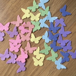 Peut inclure: Une collection de formes de papillons en feutre de différentes couleurs pastel, notamment rose, violet, bleu, vert et jaune. Les papillons sont éparpillés sur une surface en bois.