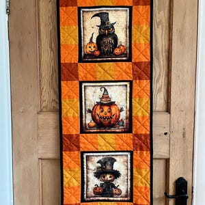 May include: A Halloween wall hanging with three square panels. The top panel depicts an owl in a witch's hat, the middle a jack-o'-lantern, and the bottom a scarecrow. The background is orange and yellow, with a black border. The hanging is on a wooden door.