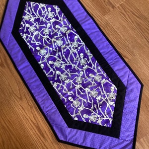 May include: Purple table runner with a black border and a white skeleton pattern on a purple background.