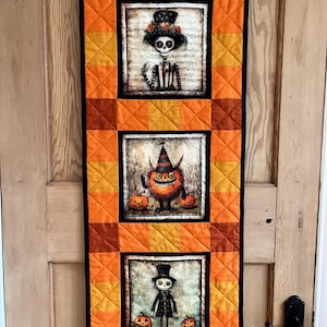 May include: A Halloween wall hanging with three framed images of quirky characters. The images are set against a quilted background of orange and yellow squares. The characters include a skeleton, a monster, and a scarecrow, each with pumpkins.