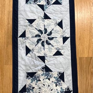 May include: A rectangular quilted table runner with a navy blue border. The runner features three floral starburst patterns in shades of blue, grey, and white. The runner is approximately 102 cm long.