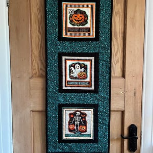 May include: A Halloween wall hanging with three square panels. The top panel shows a jack-o'-lantern and the words "FRIGHT NIGHT." The middle panel features a ghost and pumpkin with the text "A HAUNTING WE WILL GO." The bottom panel has a skeleton and pumpkins with the text "GIVE ME SOMETHING GOOD TO EAT."