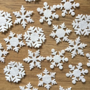 May include: A collection of white felt snowflakes of various sizes and designs. The snowflakes are scattered on a light brown wooden surface.