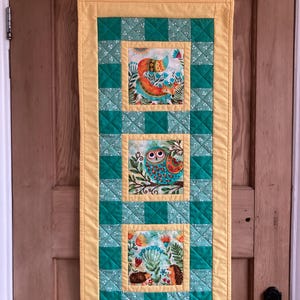 May include: A long, rectangular wall hanging with a yellow border and a green and yellow checkered pattern. The centre of the hanging features three square panels with colourful animal illustrations. The top panel shows a fox, the middle panel shows an owl, and the bottom panel shows a hedgehog.