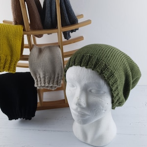 Könnte beinhalten: Eine grüne Strickmütze auf einem weißen Mannequin-Kopf. Die Mütze ist aus weichem, strukturiertem Garn gefertigt und hat eine lockere Passform.