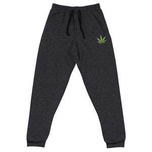 May include: Dark gray sweatpants with a black drawstring and a green cannabis leaf graphic on the left leg. The pants have a relaxed fit and cuffed ankles, suitable for casual wear.