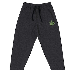 Puede incluir: Pantalones de chándal gris oscuro con cintura con cordón y un gráfico de hoja de cannabis verde en la pierna izquierda. Los pantalones tienen un ajuste relajado y tobillos con puños.