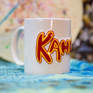 May include: White ceramic mug with the word "KACHOW" written in red and yellow letters.