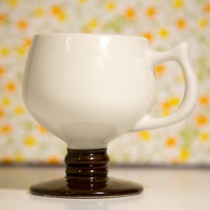 May include: White ceramic mug with a brown pedestal base and a handle. The mug is on a white surface with a floral pattern background.