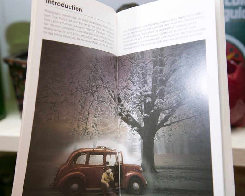 Op de afbeelding: Open boek met de titel "Introduction" zichtbaar. De pagina's tonen een foto van een vintage rode auto met een persoon, tegen een achtergrond van een besneeuwde boom en een mistig bos. De tekst op de pagina is gedeeltelijk zichtbaar.