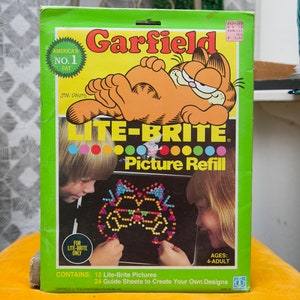 May include: A vintage Lite-Brite Picture Refill box featuring Garfield the cat. The box is green with the Garfield logo and the text "America's No. 1 Cat." It includes 12 Lite-Brite pictures and 24 guide sheets for ages 4-adult.