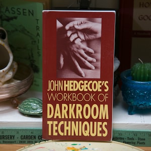Op de afbeelding: Een boek getiteld "John Hedgecoe's Workbook of Darkroom Techniques" met een bordeauxrode kaft en gouden letters. De kaft toont een zwart-wit afbeelding van handen. Andere items zijn een kikkerbeeldje en een cactuskaars.