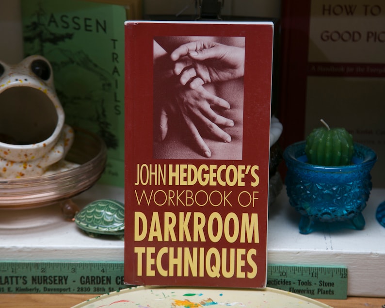 Op de afbeelding: Een boek getiteld "John Hedgecoe's Workbook of Darkroom Techniques" met een bordeauxrode kaft en gouden letters. De kaft toont een zwart-wit afbeelding van handen. Andere items zijn een kikkerbeeldje en een cactuskaars.