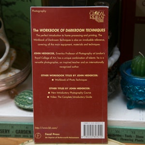 Op de afbeelding: Een rood boek met de titel "The Workbook of Darkroom Techniques" van John Hedgecoe. De tekst beschrijft het als een introductie tot thuisverwerking en afdrukken. Het boek wordt gepresenteerd op een plank met andere decoratieve items.